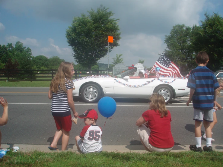 a group of children sitting on the curb with balloons near an american flag car