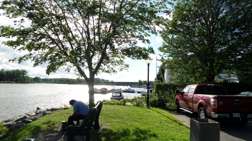 a man sits on park bench near the river while others mill about on the lawn