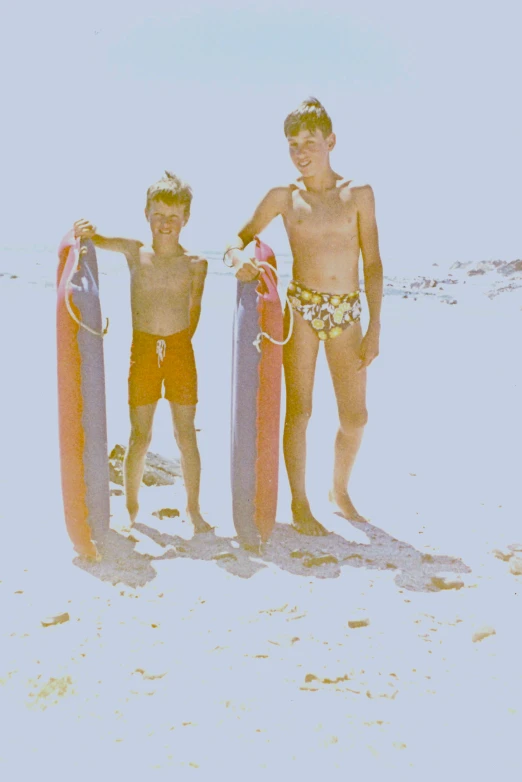 two boys in swim trunks, one holding surfboards