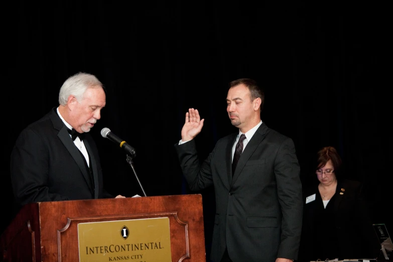 two men stand at a podium as one is holding a microphone