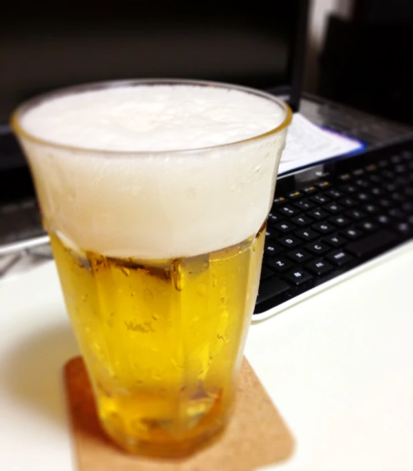 a beer in a clear glass sits on the coaster of a laptop