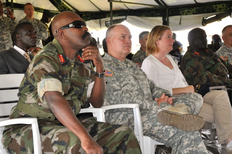 several military men sitting and standing in chairs at a meeting