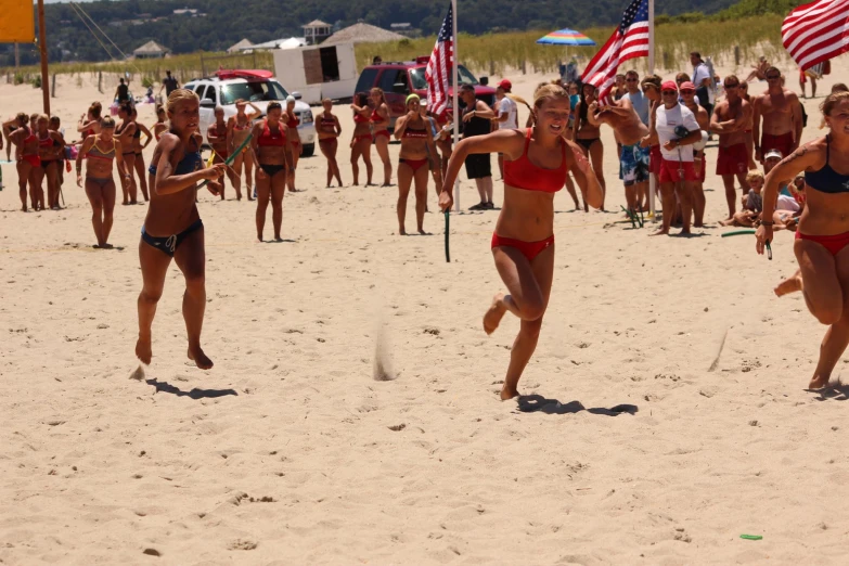 women in bikinis run across the beach as american flags are in the background