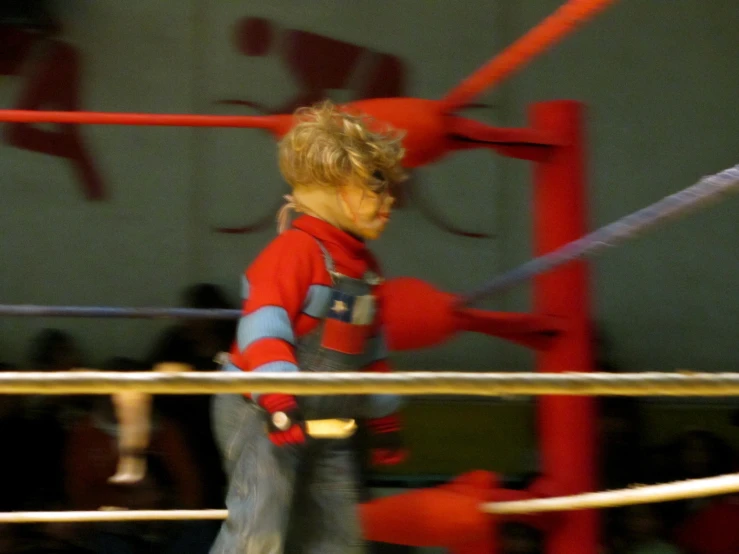 a child in a red shirt stands on one leg in front of a boxing ring and is pulling the ropes