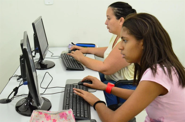 two girls sit at computers typing together