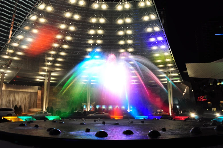 colorful fountain lit up in front of large arena with lights at night