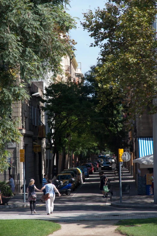 people walking down a tree lined street with cars parked on the side walk