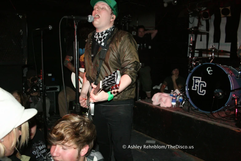 an adult with a guitar sings into microphones and some people in the audience watch