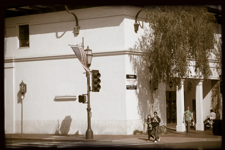 a couple walks down a city street past a tall white building with an american flag hanging on the side of it