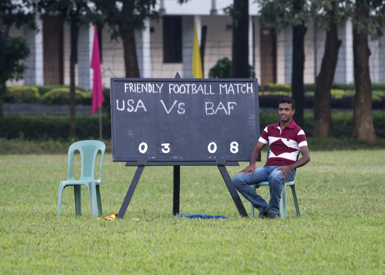 a young man sitting in a chair on the grass near a sign that says funny football match usa v bar