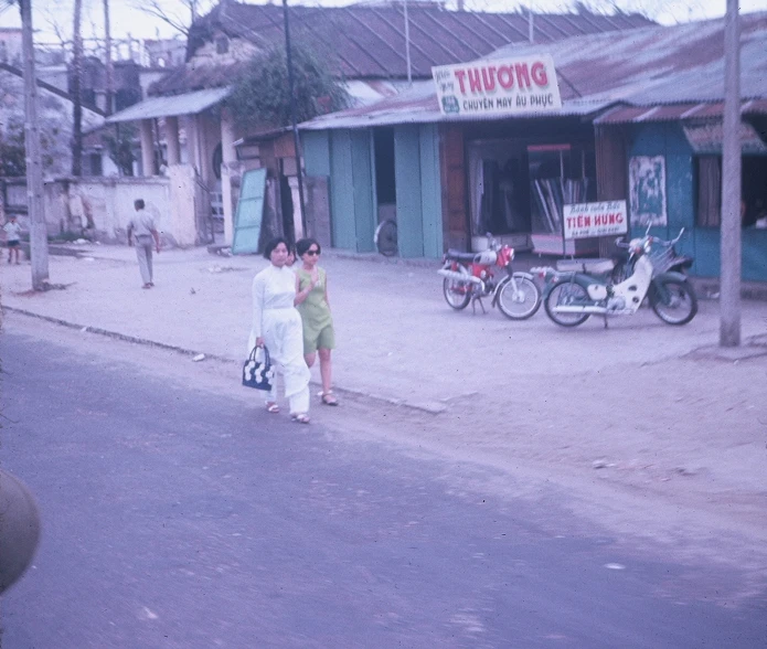 two women walking down a street with bicycles and people in the background