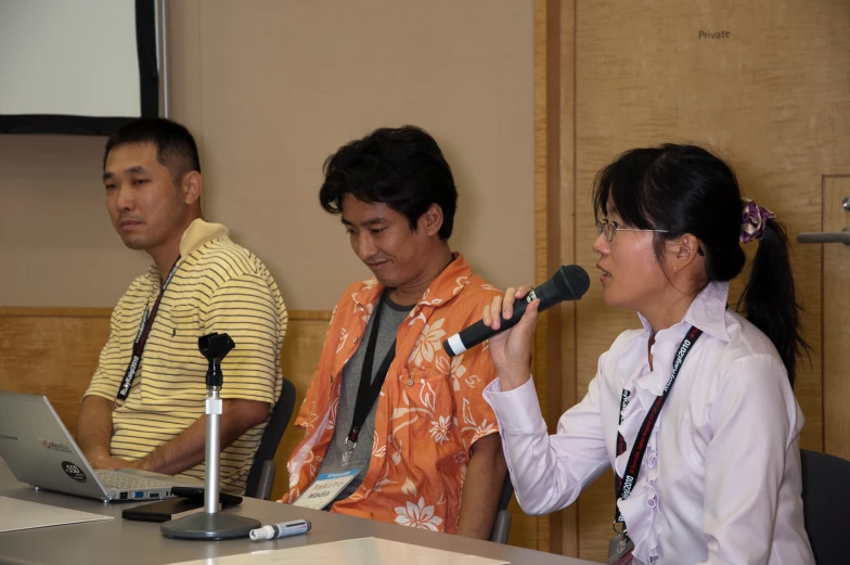 three people are sitting at a desk with microphones and one person is on a laptop