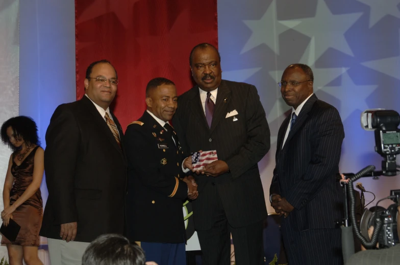 three men in suits and one woman stand beside him in front of a stage