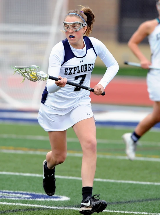 a girl wearing goggles, white uniform and a lacrosse stick running towards the camera