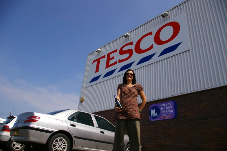 a woman in sunglasses stands near cars by a tesco sign
