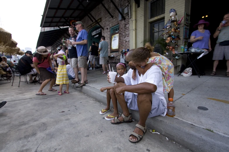 several people standing, sitting and watching two women sit on the curb and one man holds an umbrella with both hands