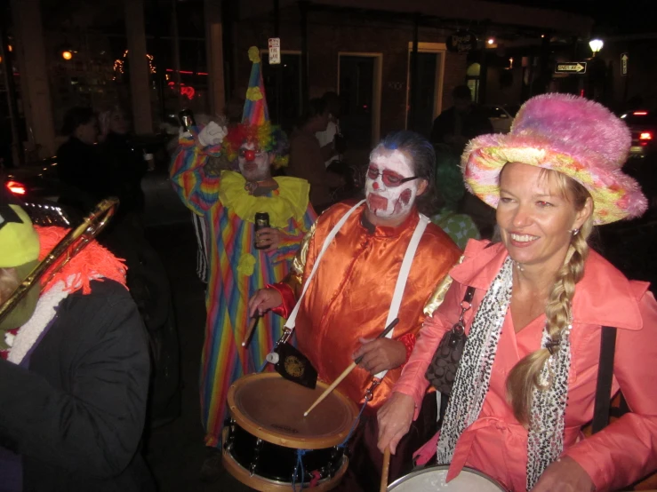 two people wearing makeup, colorful outfits, and hats, hold drums while wearing clown heads