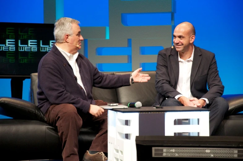 two men sit in chairs talking on a television