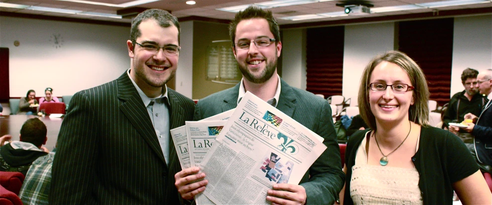 two smiling people, one holding up a paper while the other holds a phone in one hand