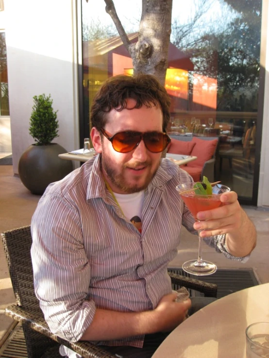 a man with sunglasses on sitting down at a table holding up a glass with a fruit in it