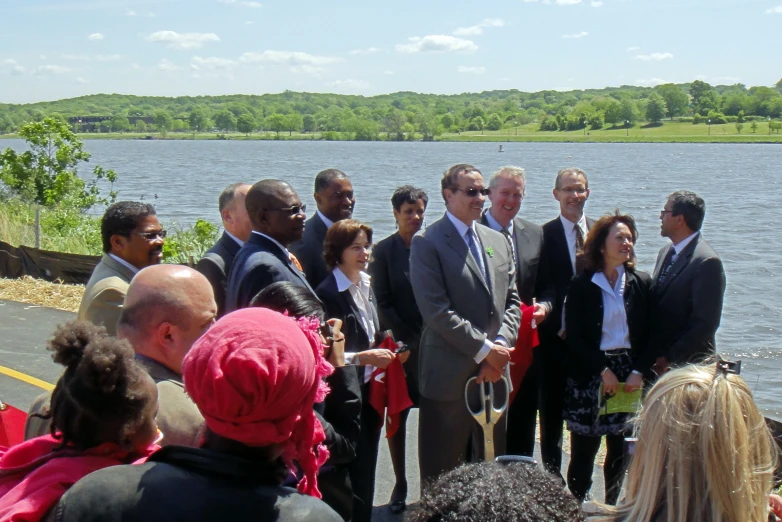 a group of people gathered at the edge of a lake with two men in suits and ties and one woman is holding scissors