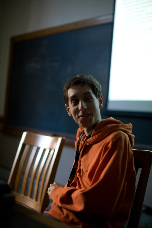 a young man sits at a table with a lecture projected behind him