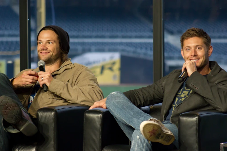 two men sitting in black leather chairs with a baseball field background