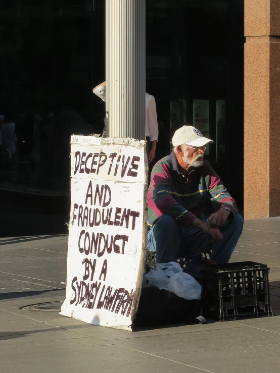 a man sits on the sidewalk next to a sign on a pole