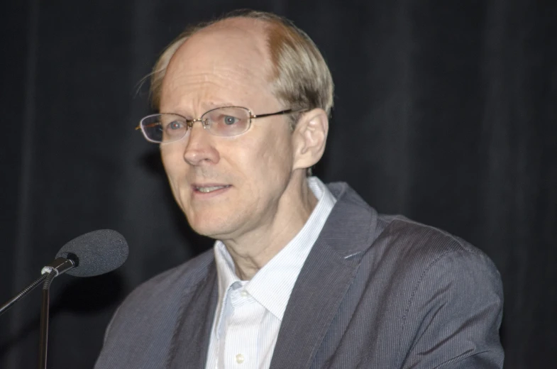 a man with glasses and a suit, making a speech at a microphone