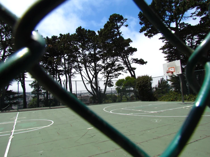 the view through a fence shows trees on a basketball court