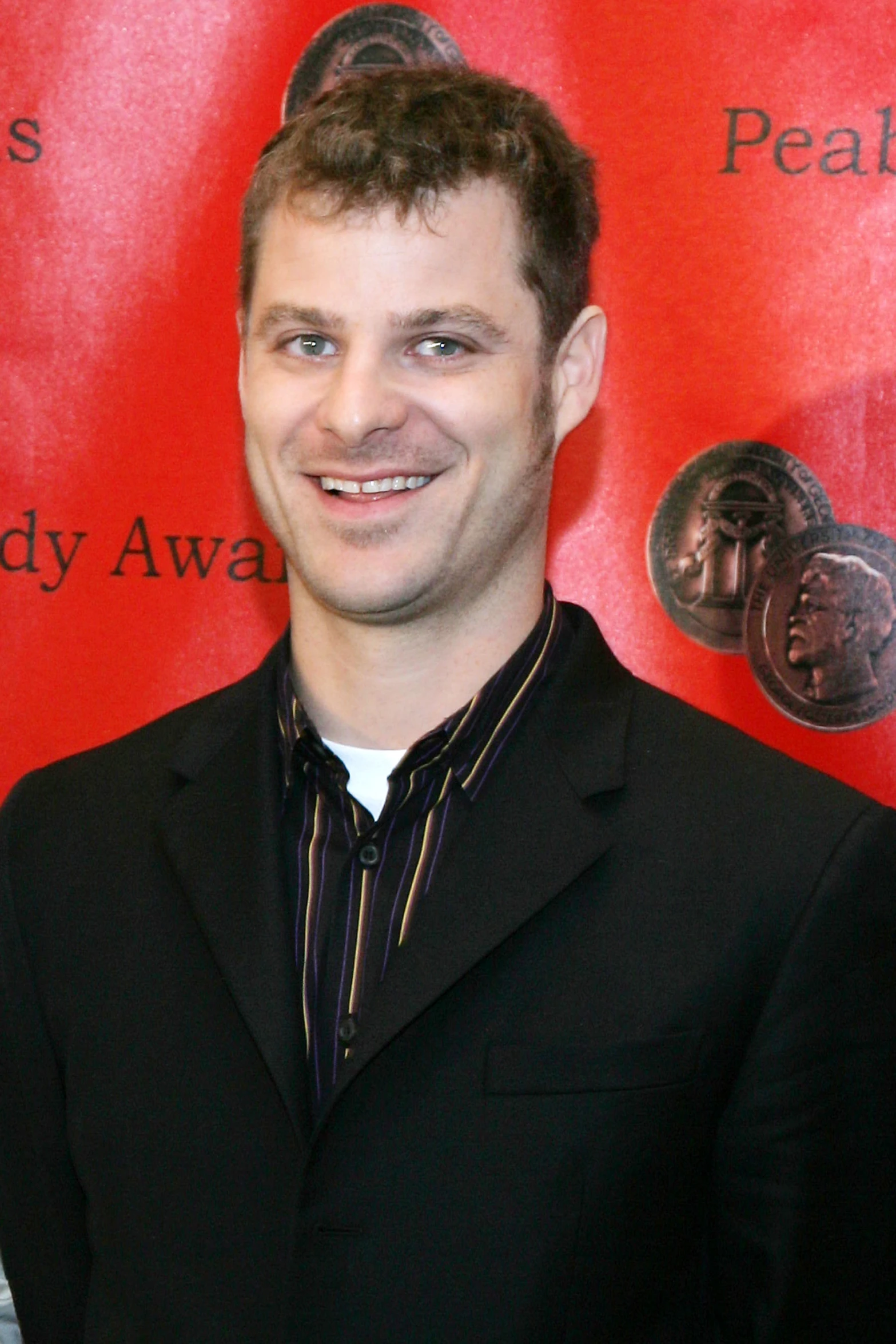 a man in a black jacket and a black and white shirt smiles at the camera