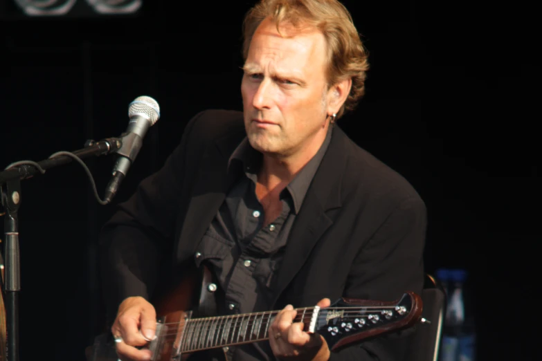 a man wearing a jacket playing guitar with a microphone behind him