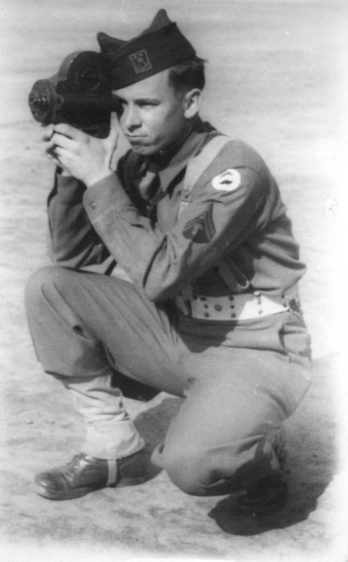 black and white pograph of boy in military uniform kneeling in the sand looking through binoculars