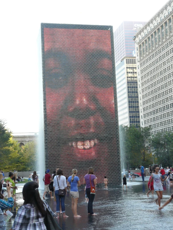 a huge face of a man with large eyes, and three fountains on the side of a city street