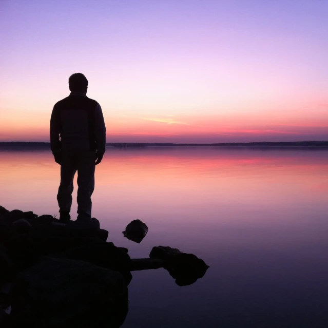man standing by the water looking at a bright purple sunset