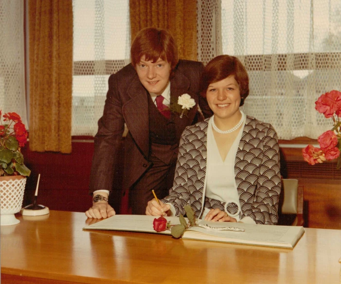 a woman poses behind a man signing a letter