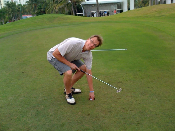 a man in gray shirt on a green putting a golf ball into a hole