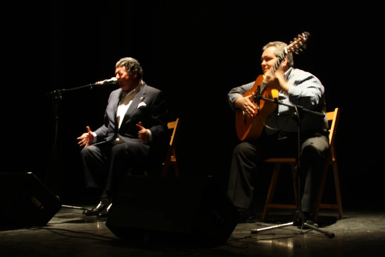 two men sit and sing together, one is playing an acoustic guitar