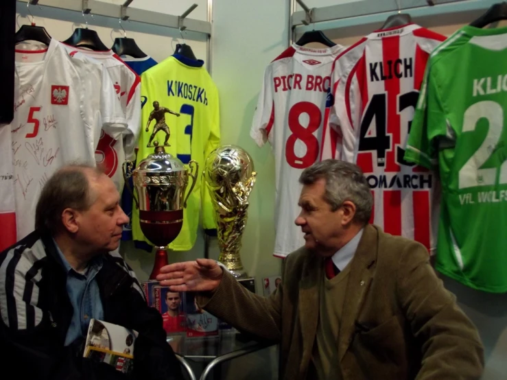 two men talking while standing in a room full of soccer jerseys