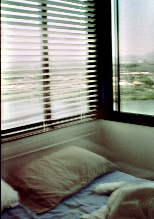 view out of a window showing a bed in a room with blinds on the windowsill