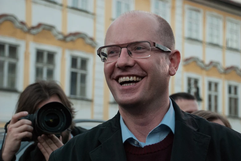 a man with glasses standing in front of a building with a camera