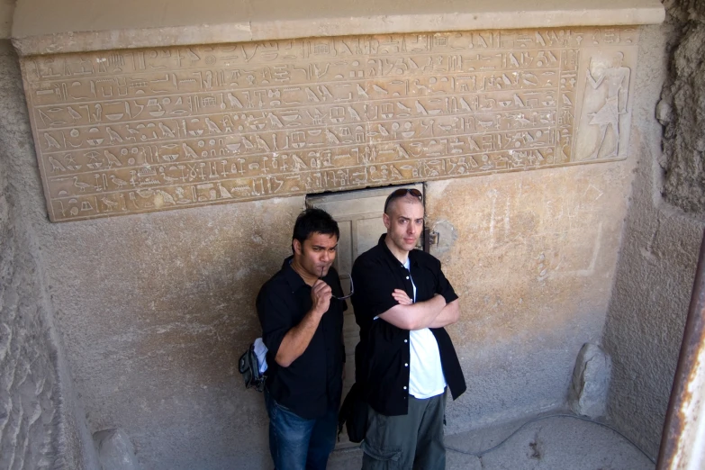 two men stand beside a stone wall with cursive writing