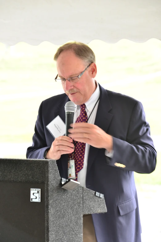 a man in suit with glasses and tie stands in front of a microphone