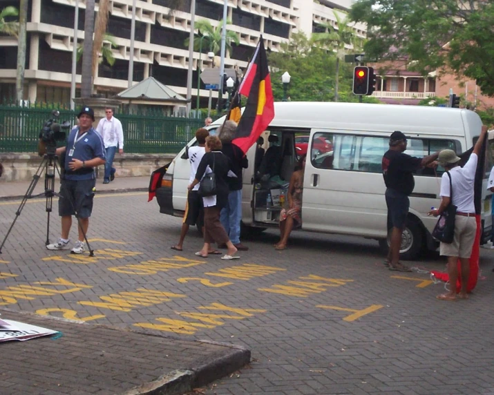 several people gathered near a small van in front of a tall building