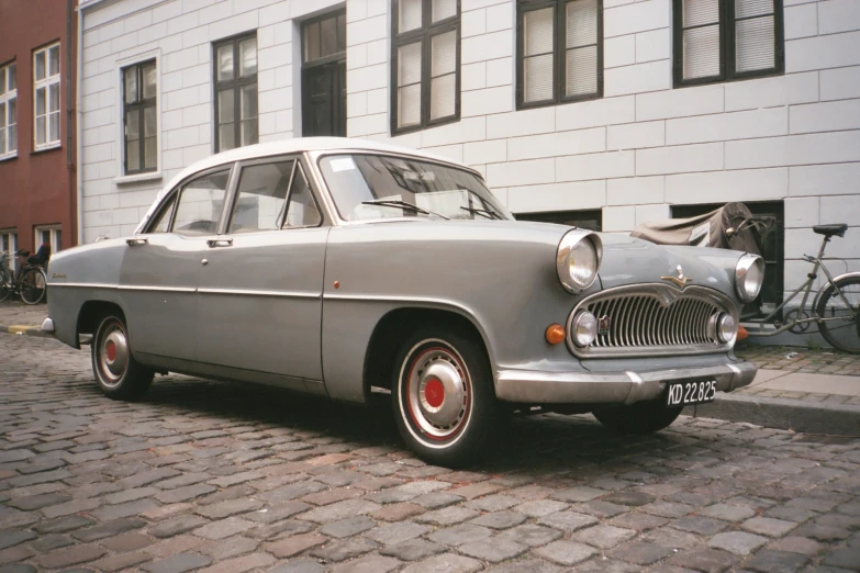 a grey classic car parked on brick road next to tall building