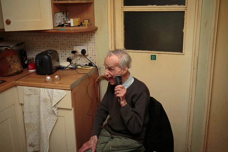 an older man sitting in his kitchen holding a microphone