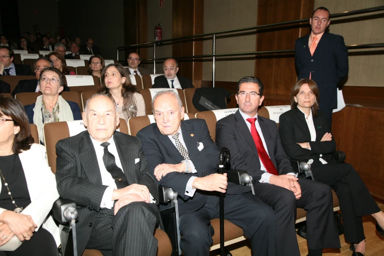 an audience of people in suits sitting in chairs and listening to a person on the stage