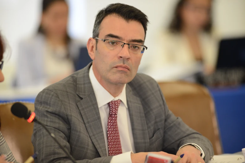 a man wearing glasses and a suit sits at a table while other people look on