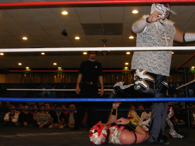 a man in the ring with a mask on and a man in a black t - shirt, standing behind the rope