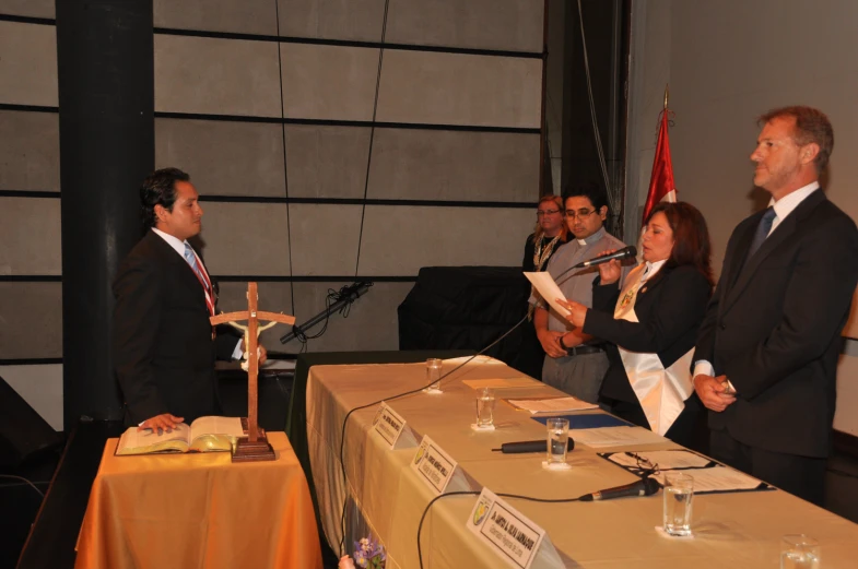 two men and one woman stand at a table with a microphone, while three other people sit down to listen